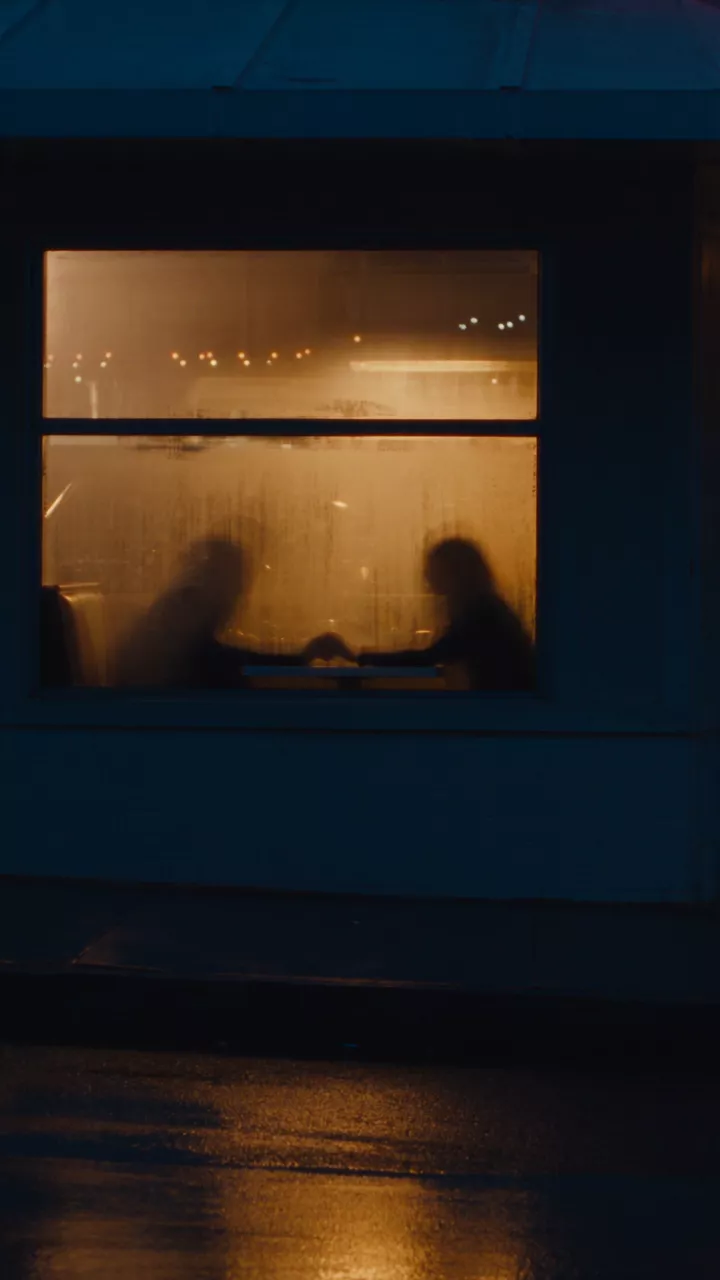 From outside at night, a glowing diner window with two blurred figures inside and its light reflected below