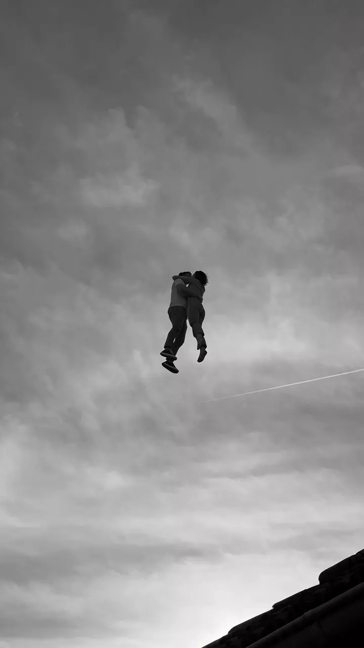 Black and white: a couple floats high among grey clouds, wrapped in a hug and kissing, with a contrail below them and a dark roof edge in the corner