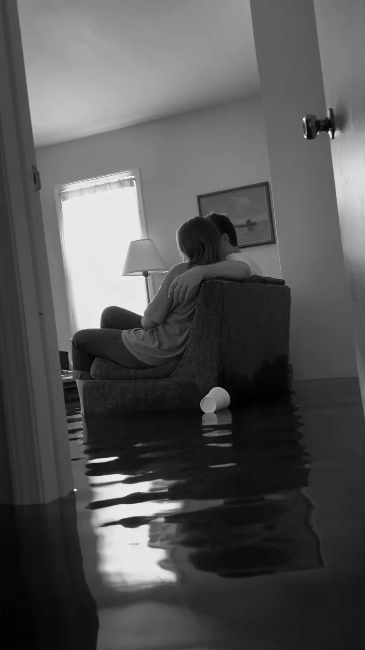 Black and white: a couple sits close on an armchair in a living room flooded with still water