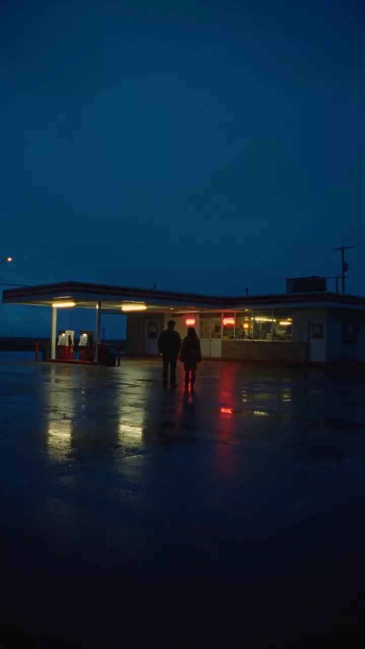 Two small figures under a lonely gas-station canopy at blue hour after rain, its lights reflected in the wet asphalt
