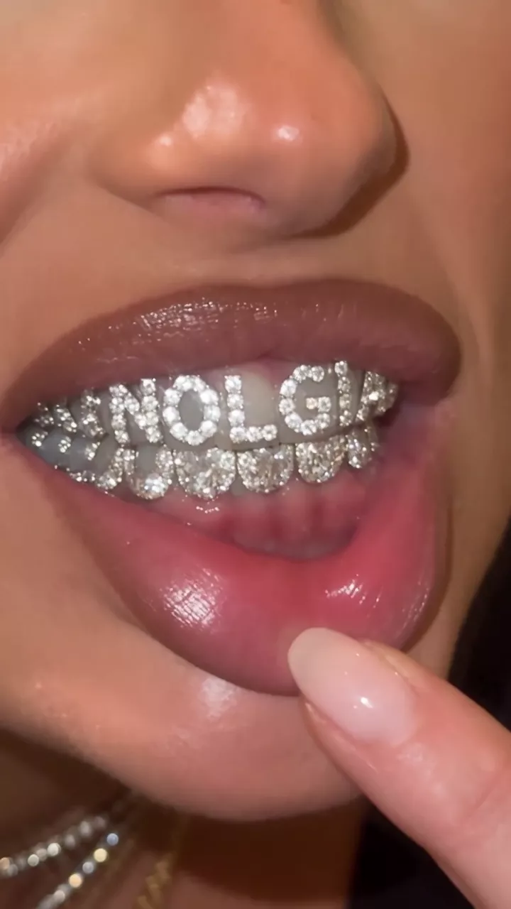 Kept: a blonde woman leans into a nightclub restroom mirror and pulls her lower lip down with one finger; the camera zooms in until a diamond grill reading NOLGIA fills the frame