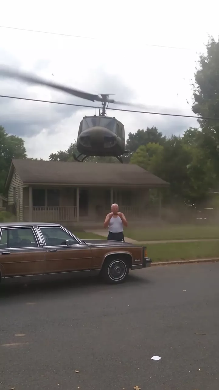 A suburban street on a cloudy afternoon: an old man in a white tank top stands by a wood-panelled sedan as a military helicopter comes in low over his house, throwing dust and paper across the street, and the camera shakes