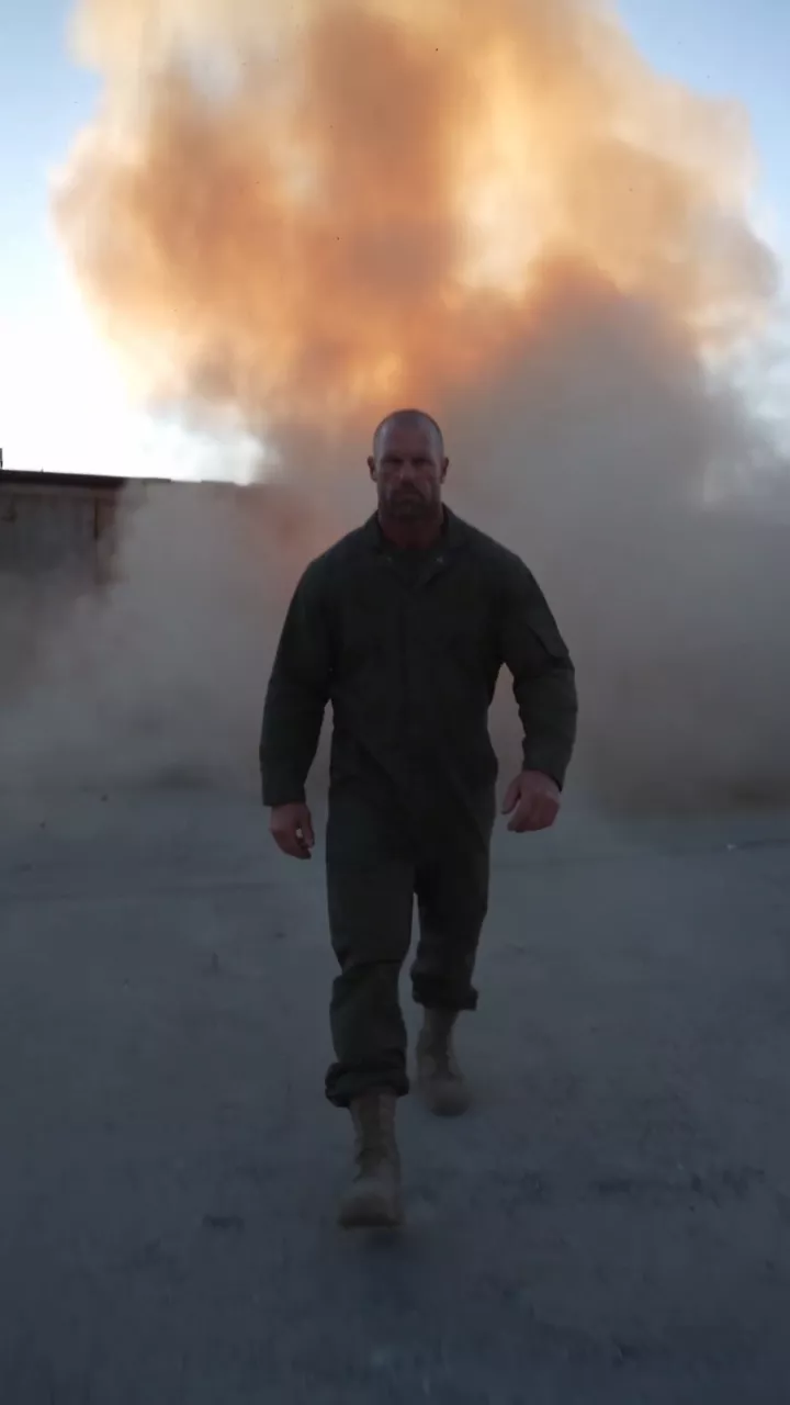An industrial parking lot at dusk: a man in dark work clothes walks toward the camera as the warehouse behind him explodes into a fireball, and he keeps walking through the smoke until he is close to the lens