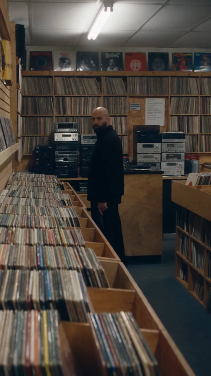 A record store after closing: a man in black stands between the record bins and looks at the camera, there is a flash, and the camera drops down behind a bin of records