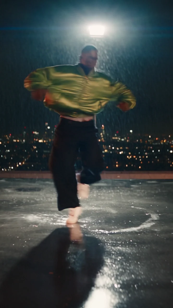 A vertical clip of the same dancer on a rain-wet rooftop helipad at night under one white spotlight, sliding low and landing in the wide stance, city lights soft behind him