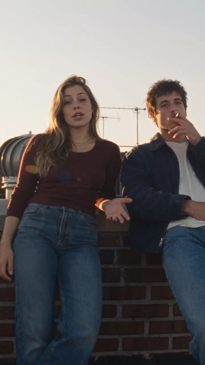 A late-afternoon New York rooftop: she leans on the brick wall talking to the camera, he sits on the parapet smoking; a fast zoom lands on her face, then a short pan finds him against the skyline