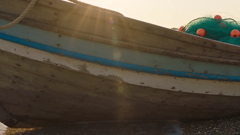 An old wooden fishing boat with peeling blue and white paint on a pebble beach at sunrise, with rope, a green net and orange floats, rendered by Seedance 2.5 at 1080p.