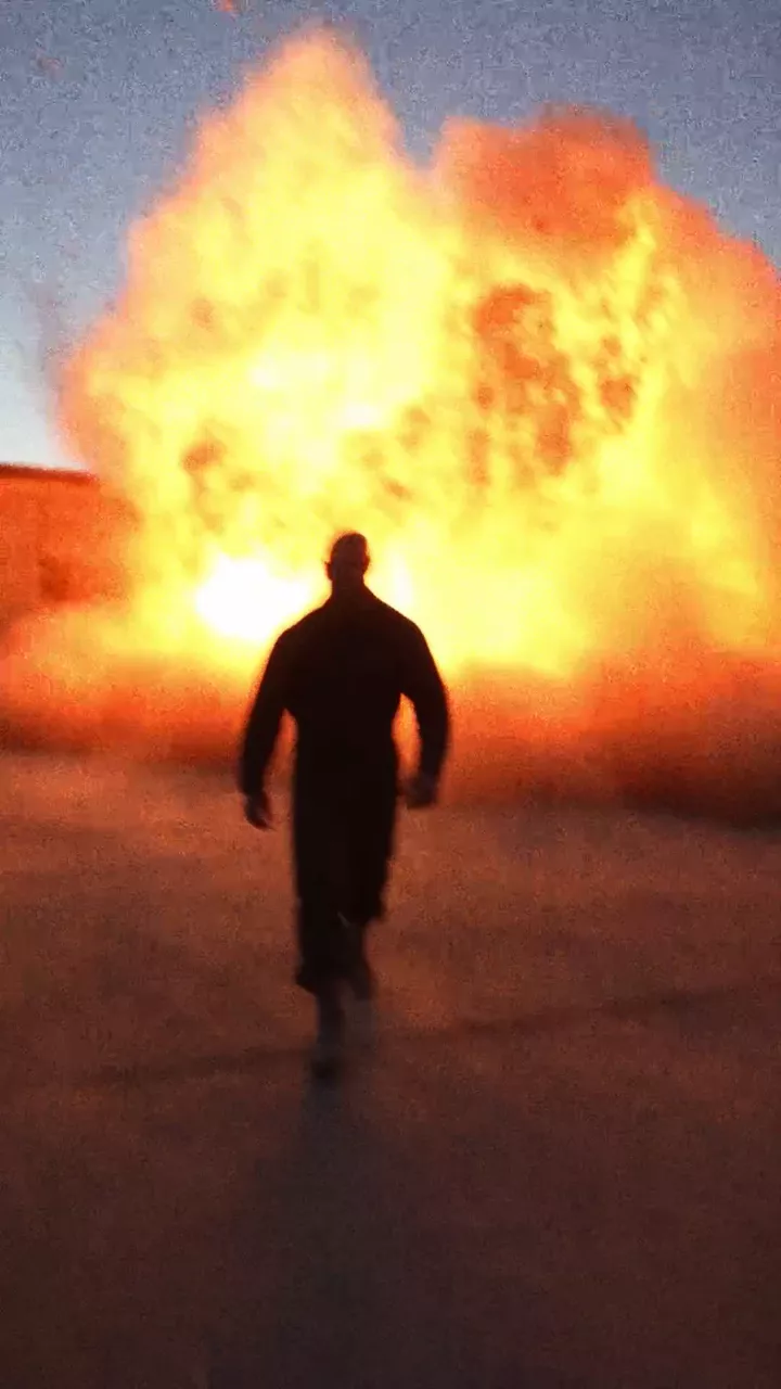 The finished Shoot out video: a man walks away from a warehouse explosion, a helicopter skims an old man's house, a man in a record store, an old man in gold chains, a man firing from a kitchen, a man kicking a car door outside a store at night, and a man in an elevator before the NOLGIA mark appears on the closed doors