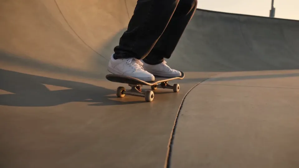 Low angle, feet only: white sneakers and black jeans on a black skateboard crossing a concrete skatepark bowl in warm evening light
