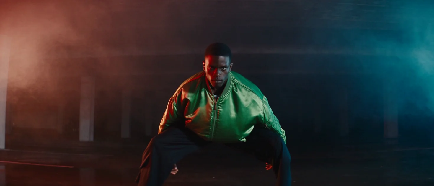 A widescreen clip of a dancer in an emerald satin bomber in a hazy underground car park lit red from the left and blue from the right, sliding low and landing in a wide stance facing the camera