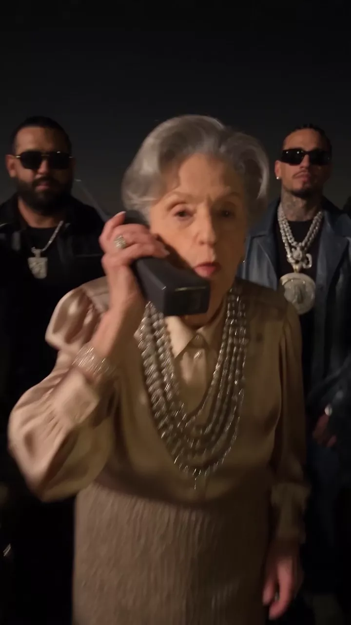 Night at a waterfront lot with a city skyline: a group in late-1990s hip-hop clothes stands by a line of sports cars, and an elderly woman in gold chains argues into a 1990s cellular phone as the camera flash fires