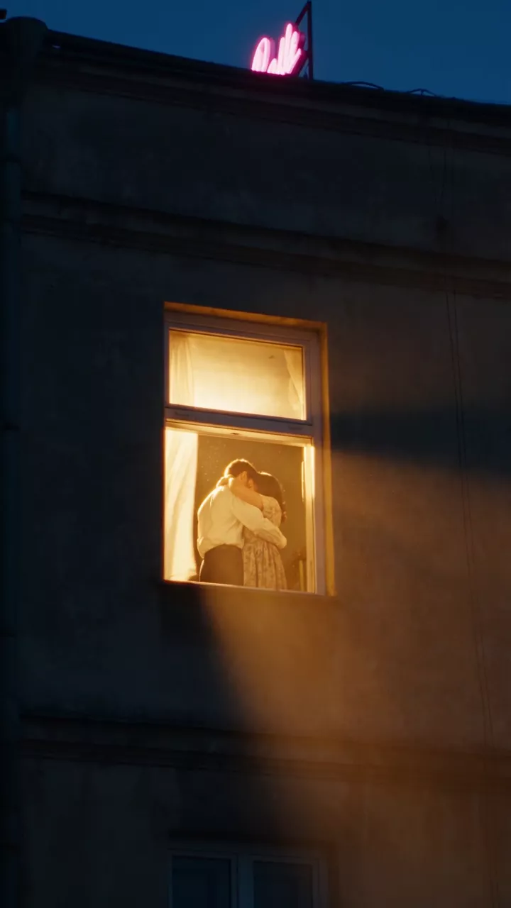 The dark side of an apartment building at night with one window glowing with warm afternoon sunlight, a couple embracing inside behind a moving curtain
