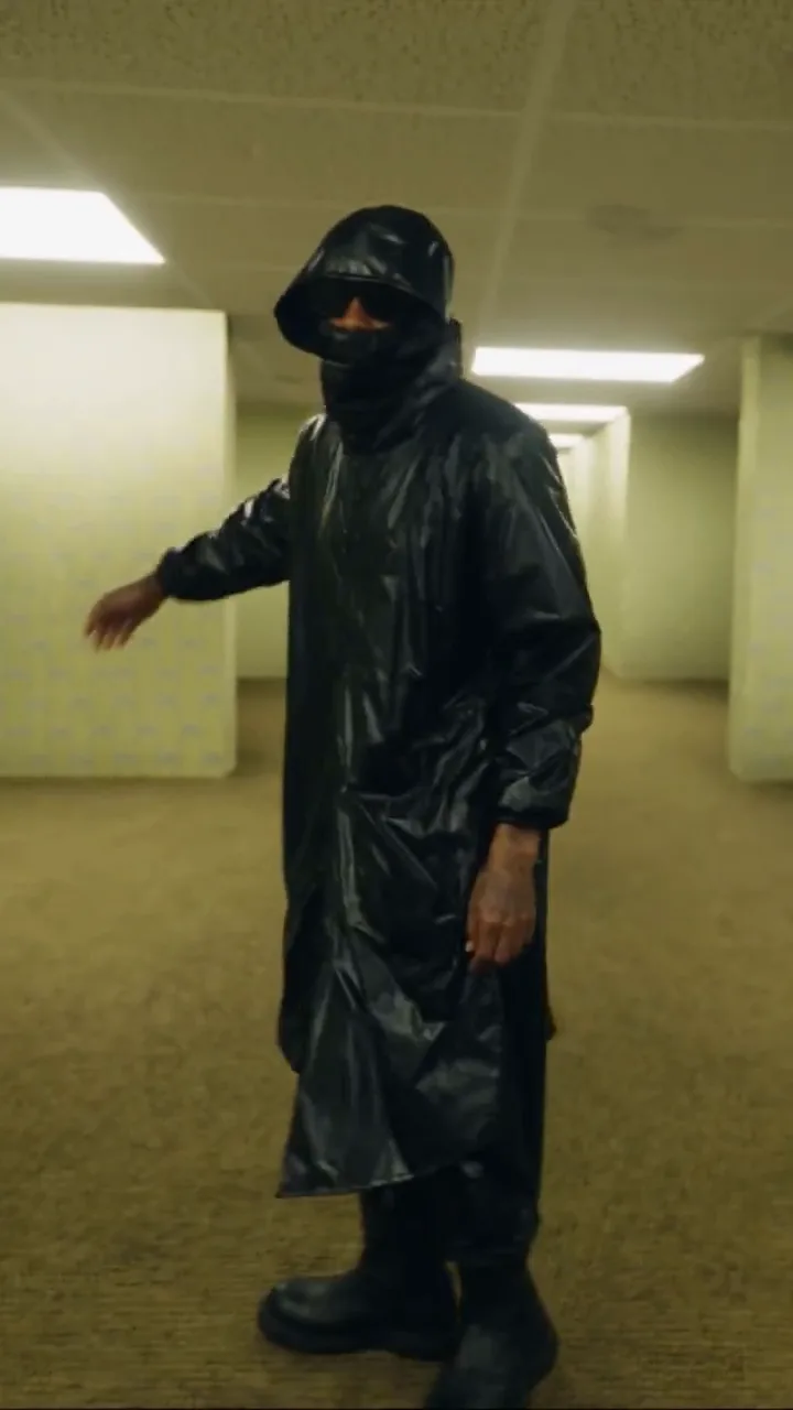 A figure in a glossy black hooded raincoat, face covering, dark glasses and chunky black boots dancing in a yellow Backrooms corridor under fluorescent panels