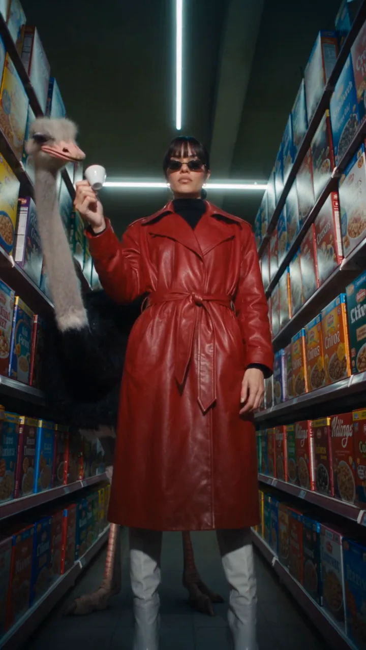 Juno in a supermarket aisle at night offering her tiny espresso to an ostrich, which takes a sip